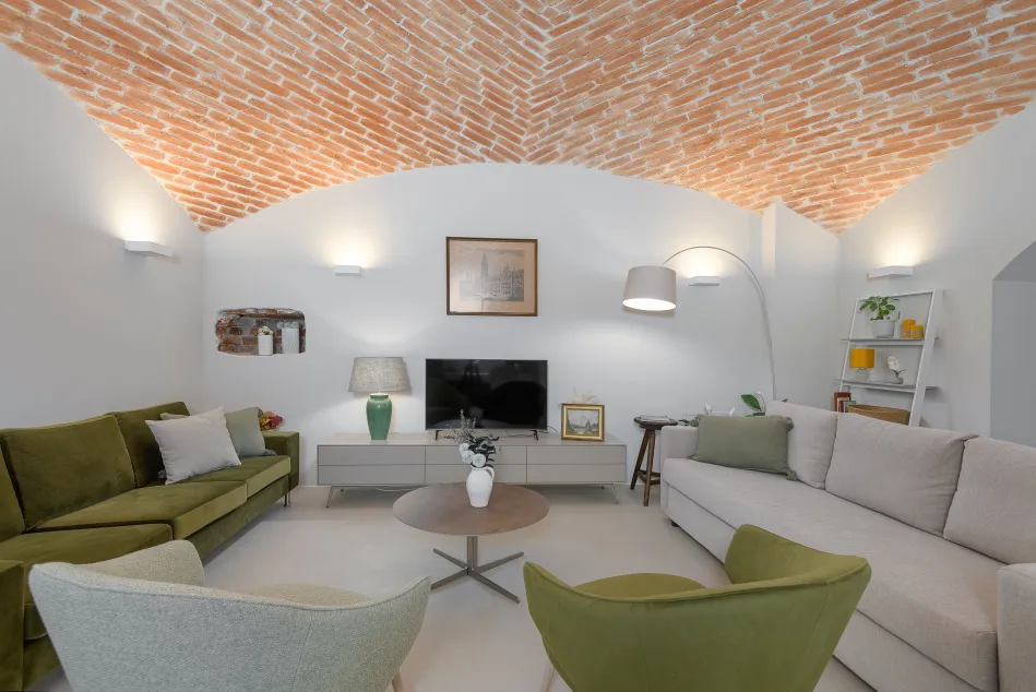 A cozy living room featuring a high, arched brick ceiling with warm lighting. The space includes a green sectional sofa with a white cushion, a beige sofa adorned with a green pillow, and two green armchairs. A round coffee table sits in the center, with a vase of flowers on it. There is a television on a light gray media console, accompanied by a decorative lamp and a green vase. A bookcase is visible in the background with plants and decorative items.