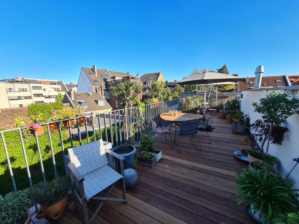 A spacious rooftop terrace in Germany featuring a wooden deck surrounded by potted plants and a small table with chairs. A large umbrella provides shade over the table, and the view includes neighboring rooftops under a clear blue sky.