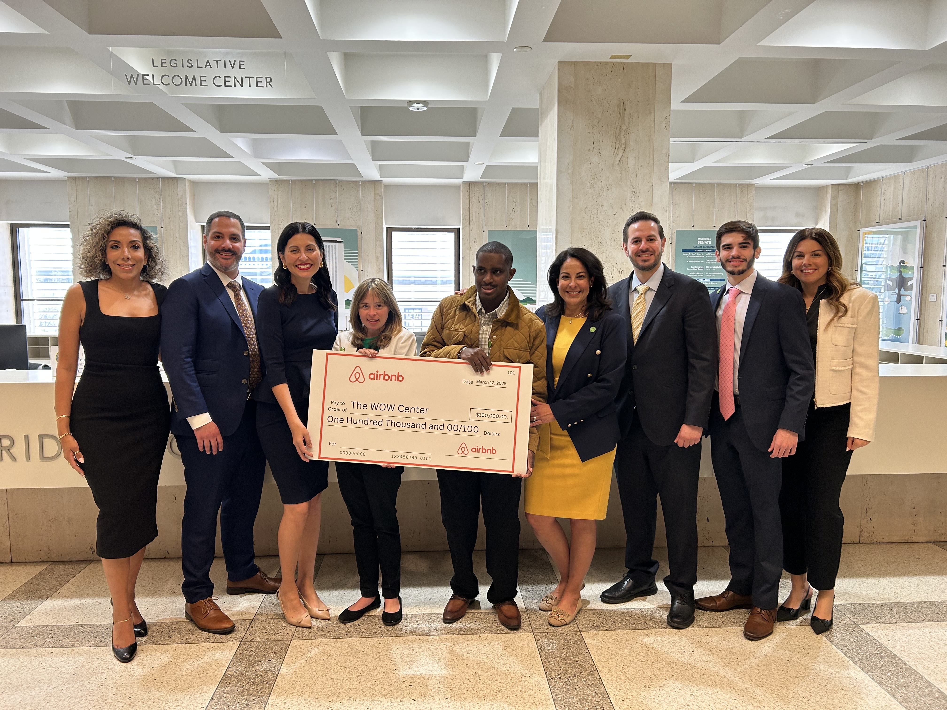 Group of nine people stand inside of a building holding a jumbo check from Airbnb addressed to The WOW Center for $100,000
