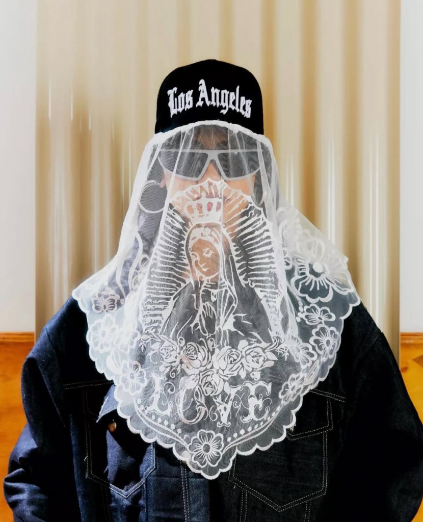 A black baseball cap with 'Los Angeles' embroidered in white, paired with a white lace veil featuring intricate floral and religious designs, worn over a dark denim jacket.