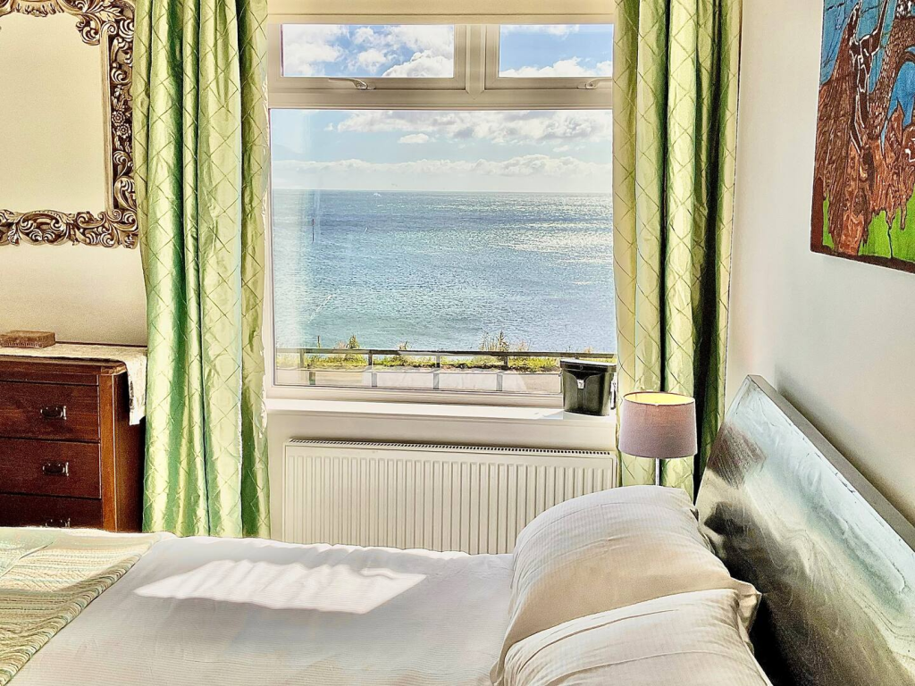Bright bedroom with large window framing a stunning ocean view under a sunny sky, green curtains, and cozy furnishings.