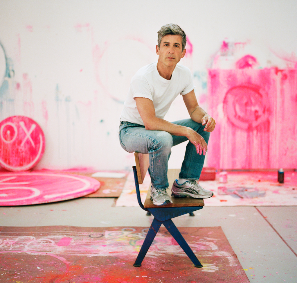 A casually dressed individual sits on a modern stool surrounded by a studio filled with pink art and paint splatters.